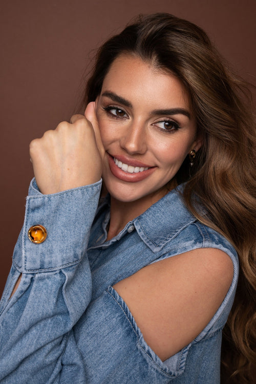 Portrait of a woman wearing a light denim off-shoulder blouse with a black and goldbrown button cover detail on the sleeve.