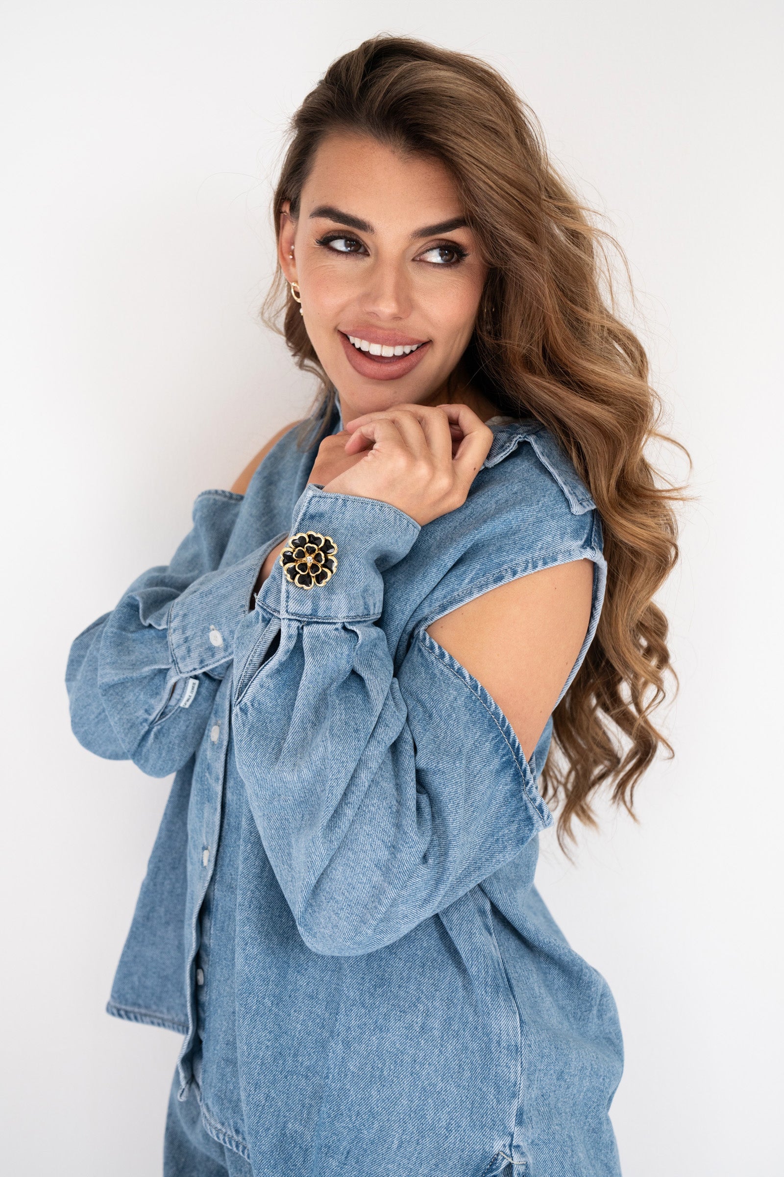 Woman wearing a light blue denim blouse with shoulder cut-outs, accessorized with a black and gold statement button cover on the cuff for a chic, elevated look.
