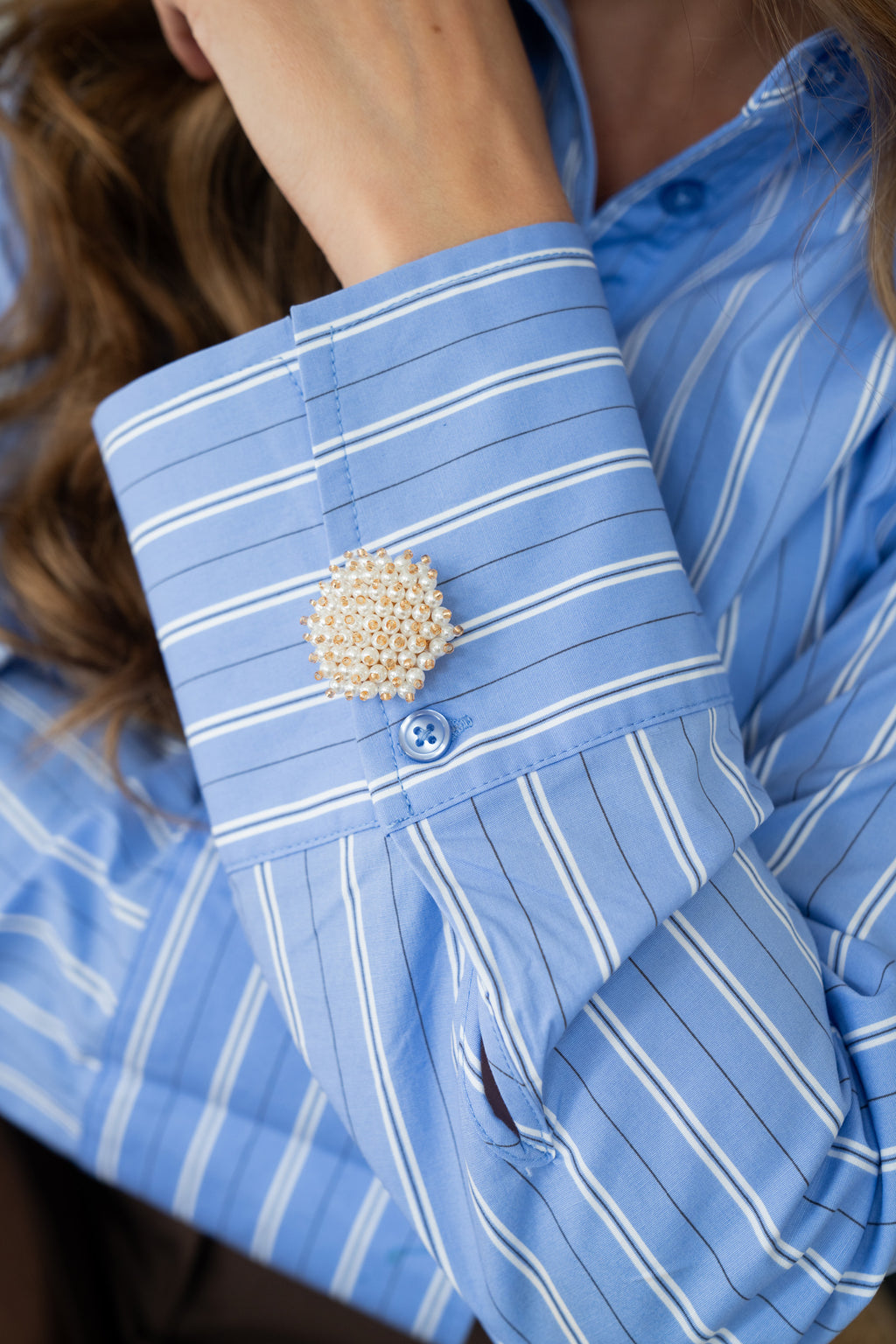Close-up of a blue striped shirt cuff styled with a pearl and gold button cover, adding an elegant statement detail to a casual outfit.