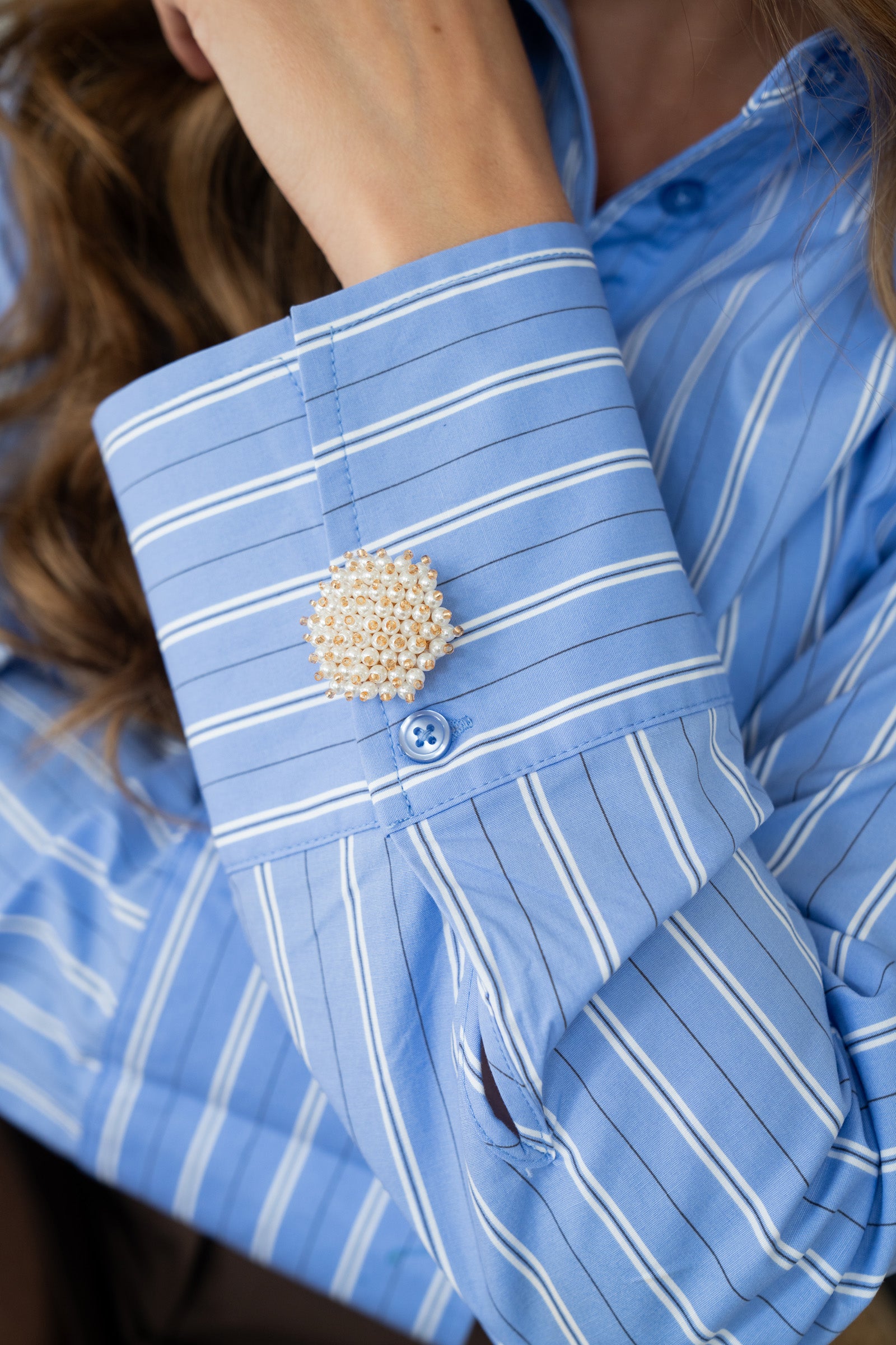 Close-up of a blue striped shirt cuff styled with a pearl and gold button cover, adding an elegant statement detail to a casual outfit.